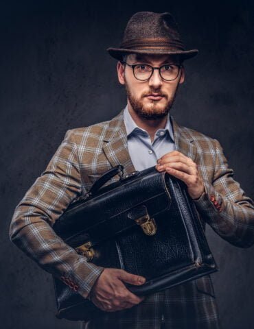 bearded man wearing casual suit hat glasses holds sui scaled